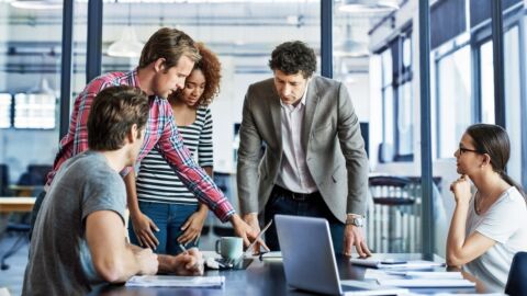 Eine Meeting-Situation mit fünf Personen, die um einen Tisch herum sitzen und stehen.