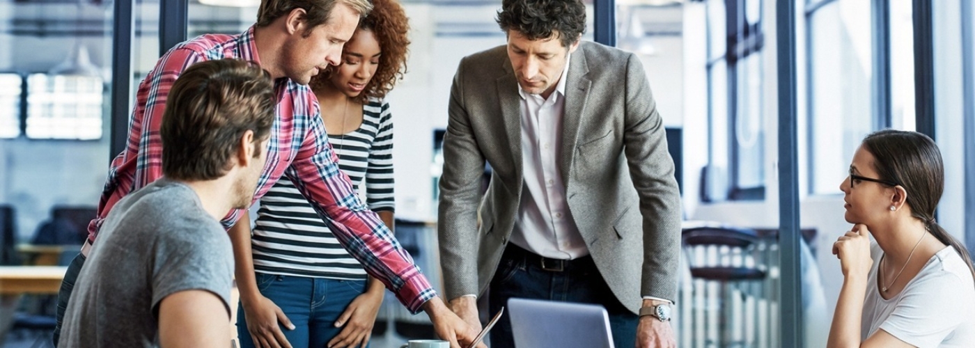 Eine Meeting-Situation mit fünf Personen, die um einen Tisch herum sitzen und stehen.