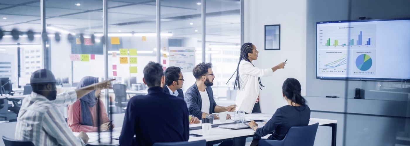 Eine Gruppe Menschen sitzt an einem Besprechungstisch und diskutieren eine Grafik, die auf einem Monitor eingeblendet ist.