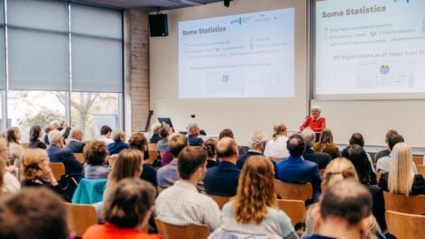 Veranstaltungsraum mit vielen Menschen, die auf Stühlen sitzen und zu einer Rednerin nach vorne schauen.