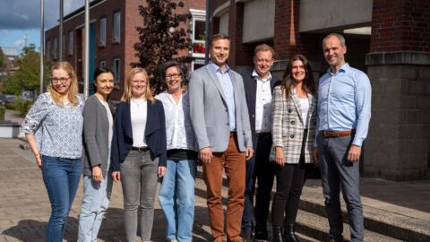 Acht Personen stehen nebeneinander vor einem Bürogebäude und lächeln in die Kamera.
