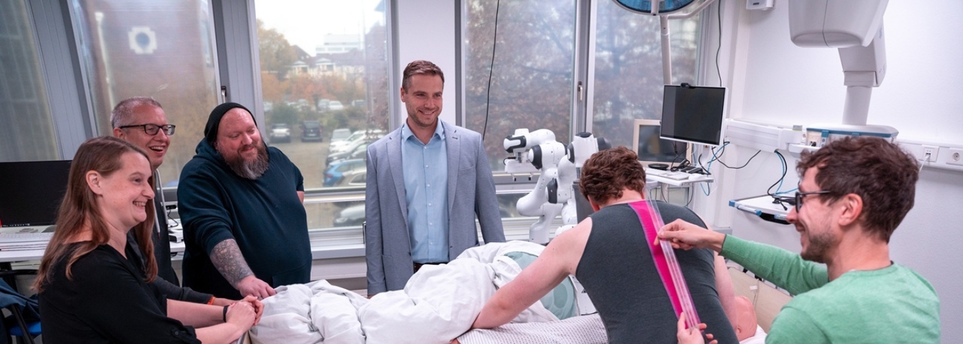 Eine kleine Gruppe von Menschen steht an einem Pflegebett, in dem eine Puppe liegt. Ein Mann dreht diese Puppe, während ein anderer Mann einen Sensorstreifen an seinen Rücken hält.