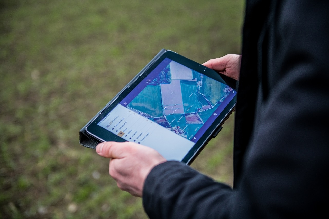 Eine Person hält ein Tablet fest und steht auf einem Acker