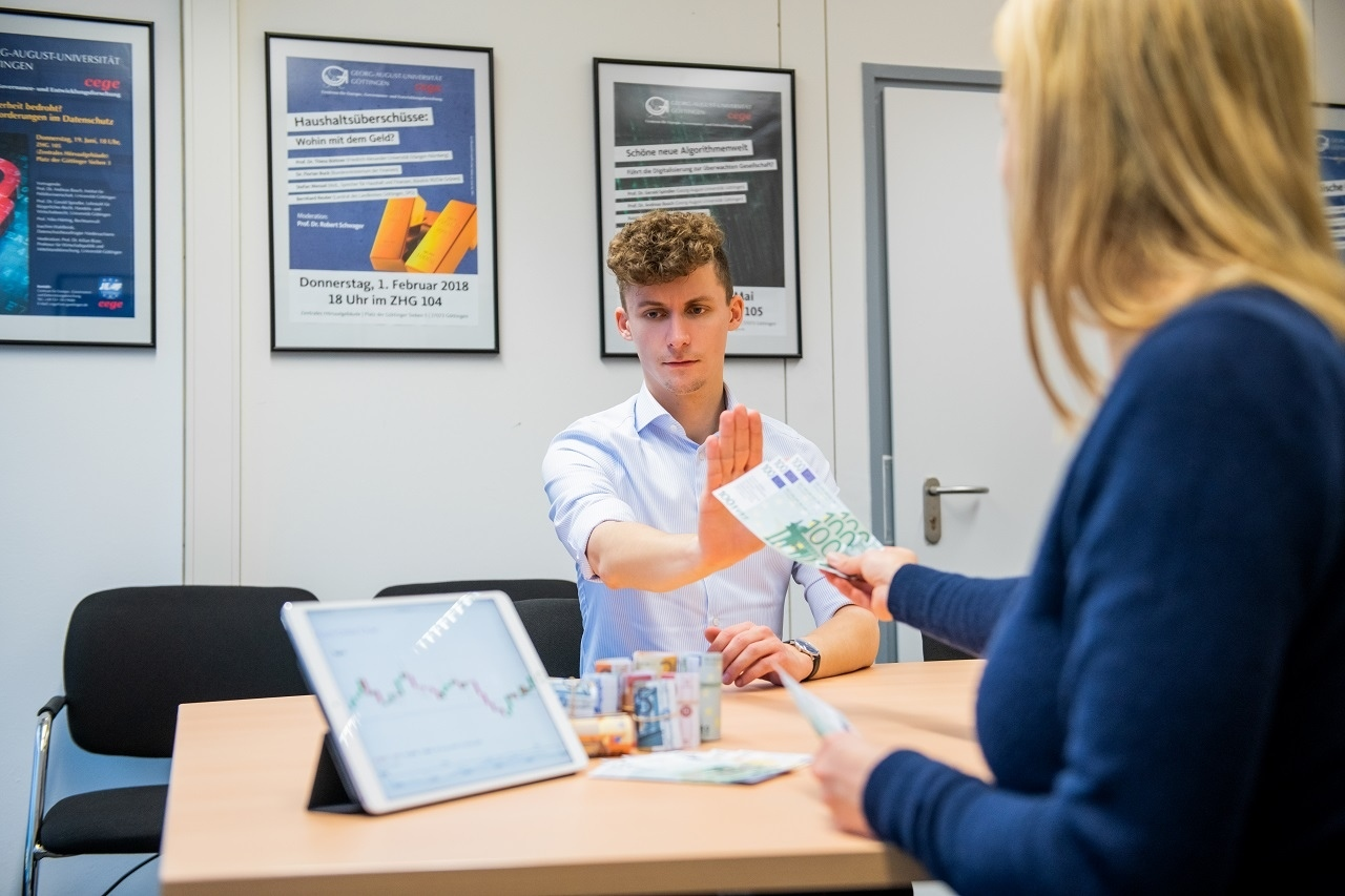 Ein Mann lehnt Geld von einer Frau ab, während beide an einem Tisch sitzen.