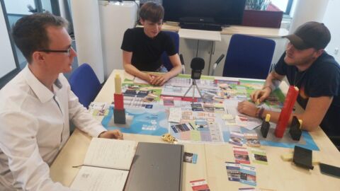 Drei Personen sitzen mit einem Spiel und Mikrofon am Tisch