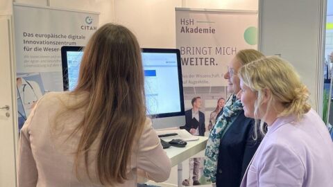 Drei Frauen stehen vor einem Bildschirm.