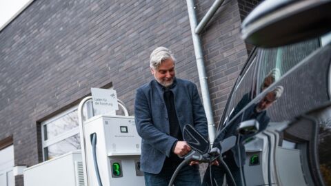 Mann steht an E-Ladesäule und lädt ein Auto auf