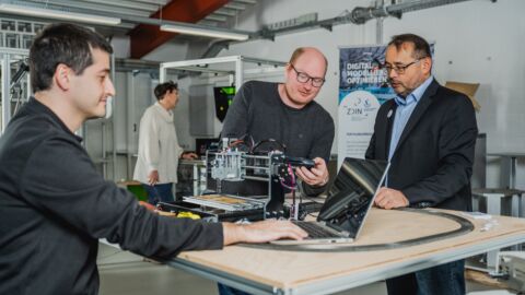 Drei Männer sind an einem Tisch auf dem eine Graviermaschine steht