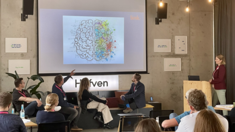 Ein Publikum hört einer Präsentation zu. Auf der Leinwand sind zwei gezeichnete Gehirnhälften.