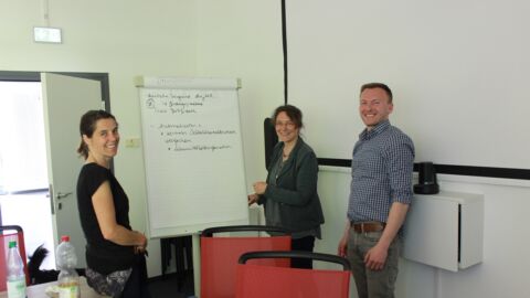 Zwei Frauen und ein Mann stehen vor einem Flipchart in einem Meetingraum.