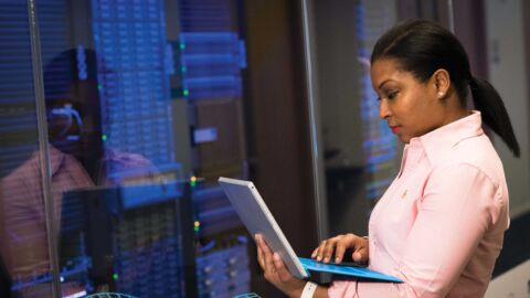 Frau steht in einem Serverraum und blickt auf einen Laptop