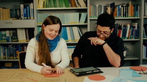 Zwei Personen sitzen an einem Tisch nebeneinander und reden miteinander