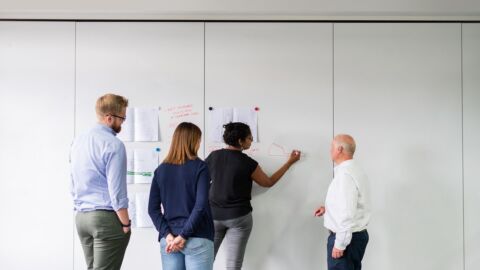Vier Personen stehen vor einer Wand an der Zettel mit Magneten befestigt sind.