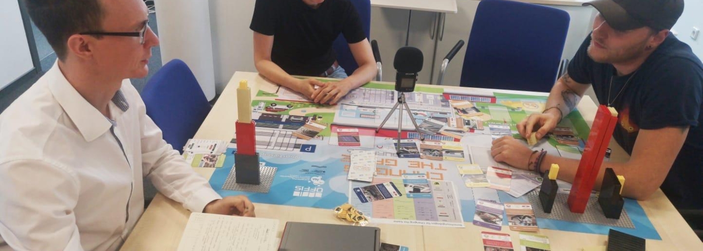 Drei Personen sitzen mit einem Spiel und Mikrofon am Tisch