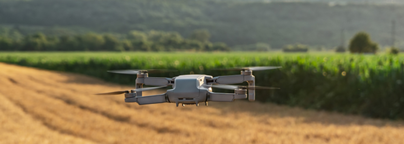 Eine Drohne fliegt über einem Feld, ein Wals ist im Hintergrund