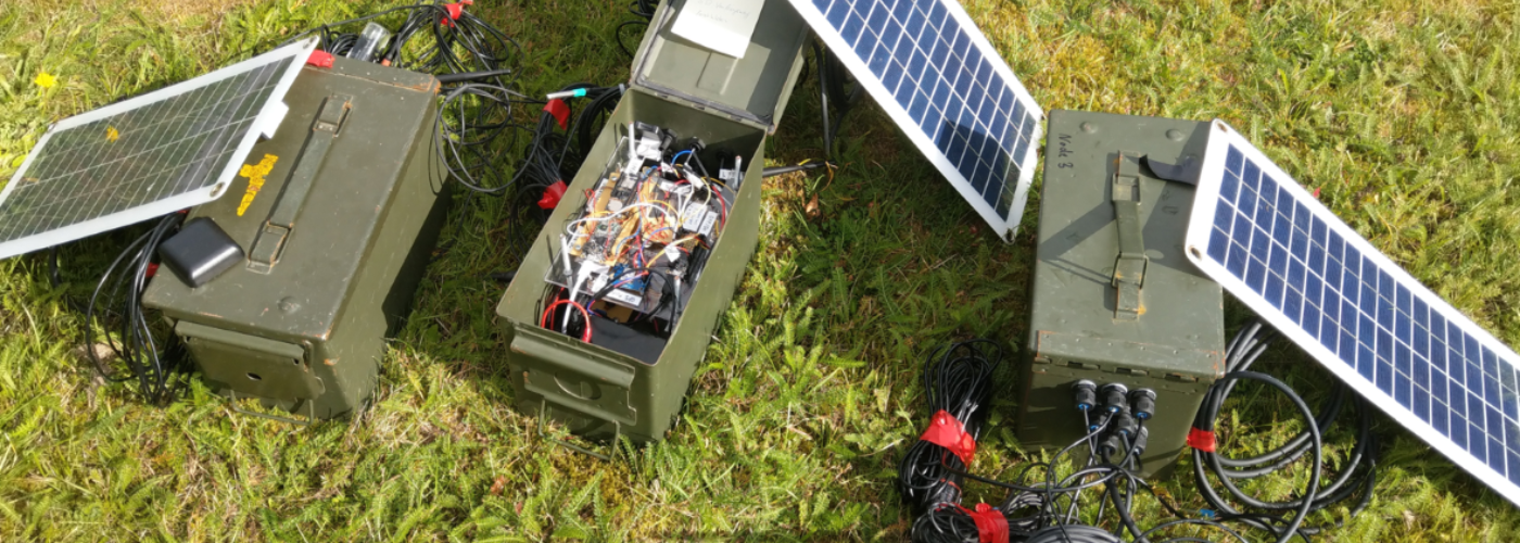 Auf dem Bild sind grüne Metallkisten zusehen, die mit Kabeln und Technik gefüllt sind. Außerdem sind Solarpanele zusehen.