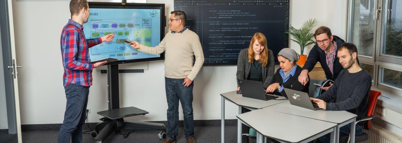 Mehrere Personen in einem Raum arbeiten an einem Bildschirm und an einem Tisch mit Laptops