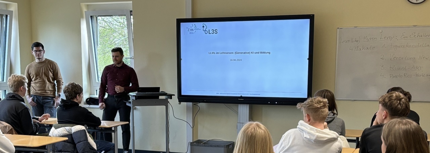 Zwei Männer halten in einem Klassenraum eine Präsentation. Schüler hören zu.