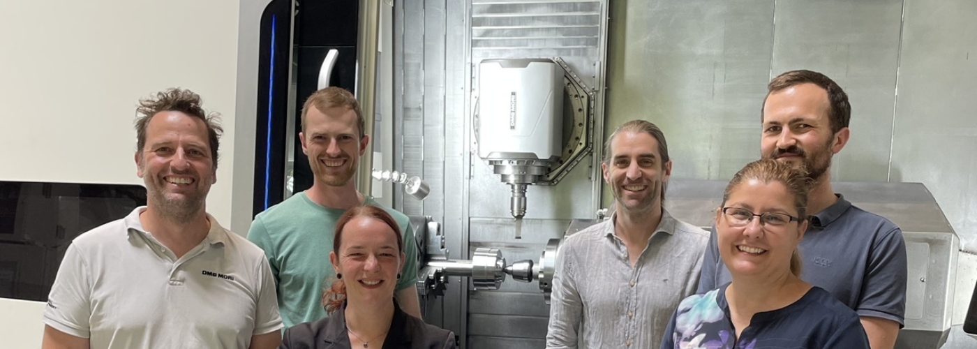 Vier Männer und zwei Frauen stehen vor einer Produktionsanlage und lächeln in die Kamera.