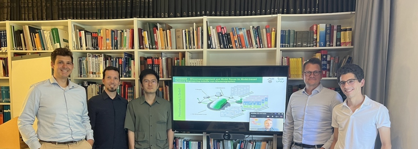 Fünf Männer stehen in einem Meetingraum, im Hintergrund ist eine Regal mit Büchern zu sehen, davor ein Monitor mit einer Grafik.