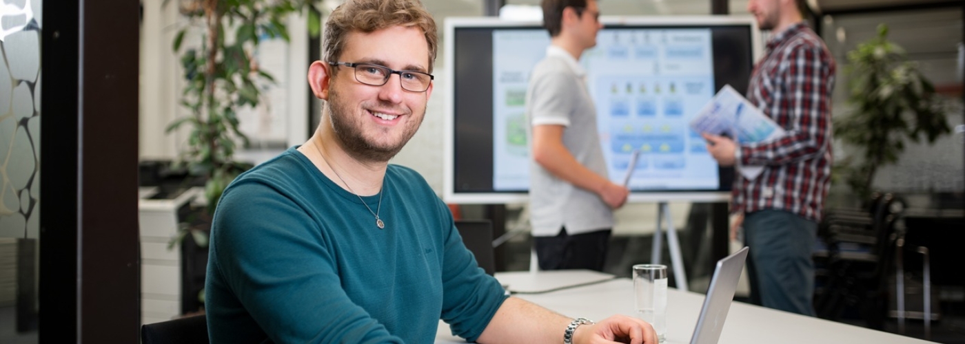 Ein Mann sitzt an einem Tisch mit geöffnetem Laptop und lächelt in die Kamera. Im Hintergrund stehen zwei Männer voreinander und unterhalten sich.