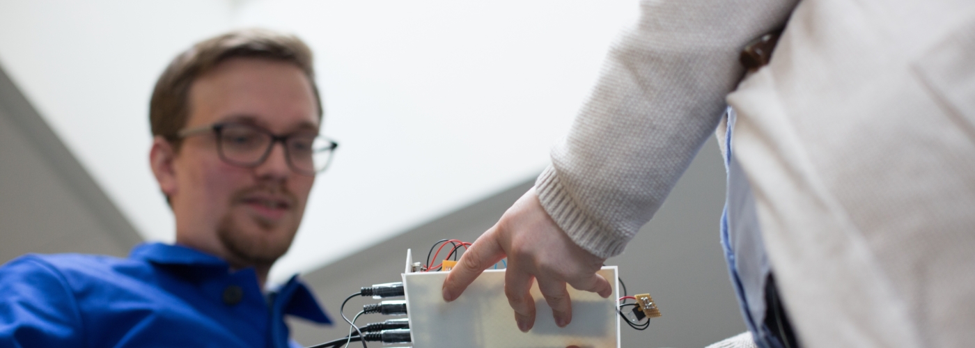 Frau hält Platte mit Kabeln, auf die ein Mann blickt
