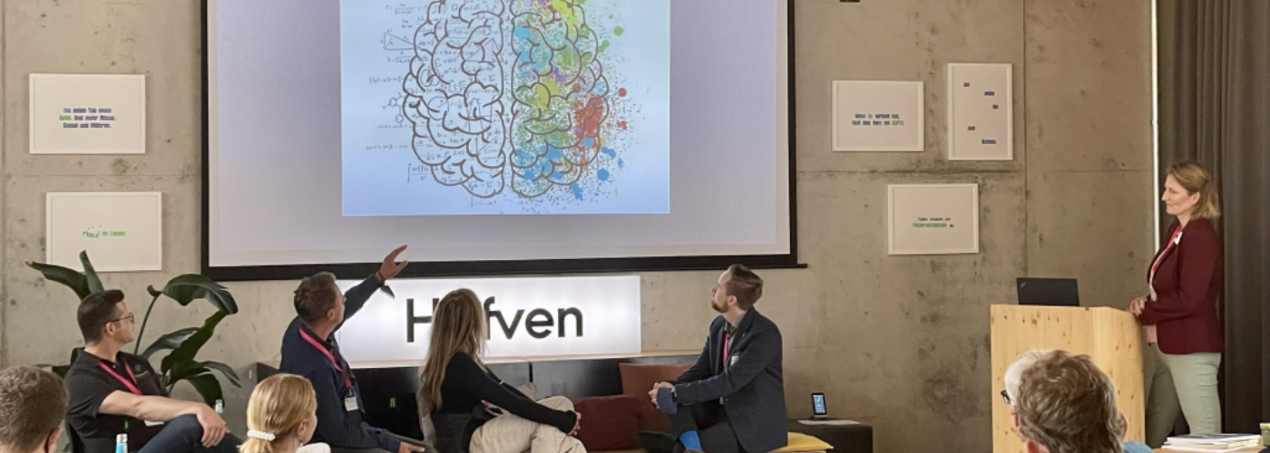 Ein Publikum hört einer Präsentation zu. Auf der Leinwand sind zwei gezeichnete Gehirnhälften.