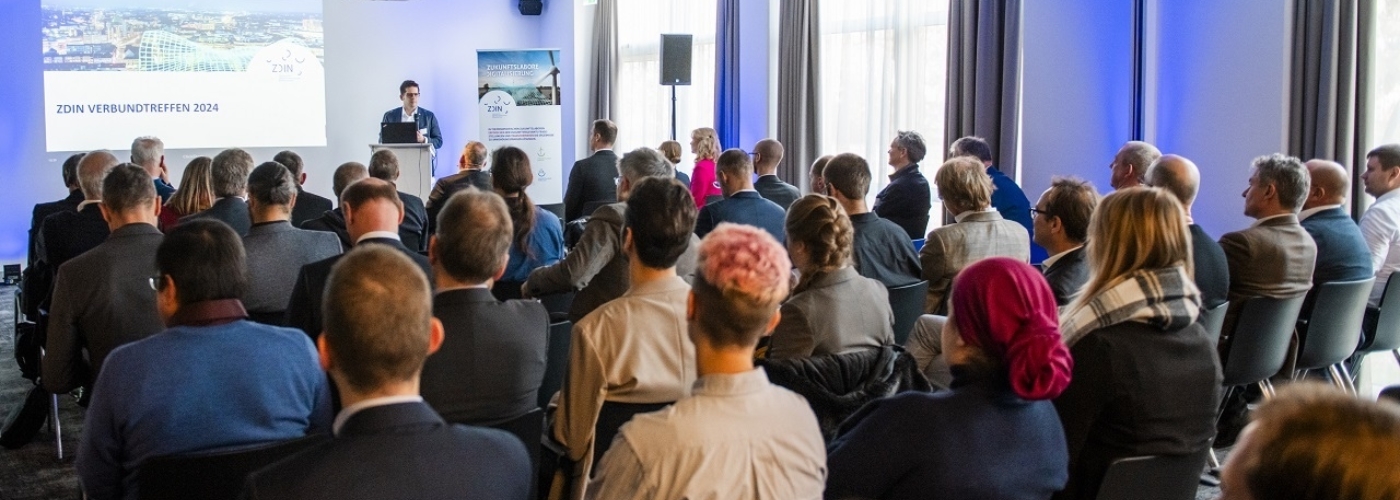 Viele Menschen sitzen in einem Raum und schauen auf eine Leinwand, vor der ein Mann am Rednerpult steht.