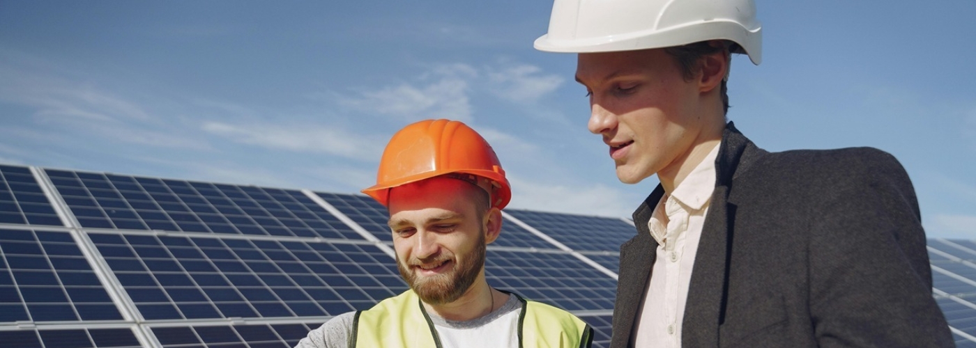 Zwei Männer mit Bauhelmen und einem Plan stehen vor einem Solarfeld.
