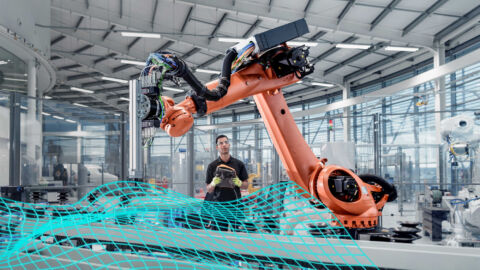 Ein Mann steht mit einer Steuerung in einer Produktionshalle vor einer Maschine mit einem Roboterarm.