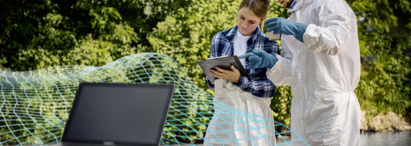 Eine Frau und ein Mann stehen mit einem Laptop, einem Klemmbrett und einer Wasserprobe an einem Fluss.